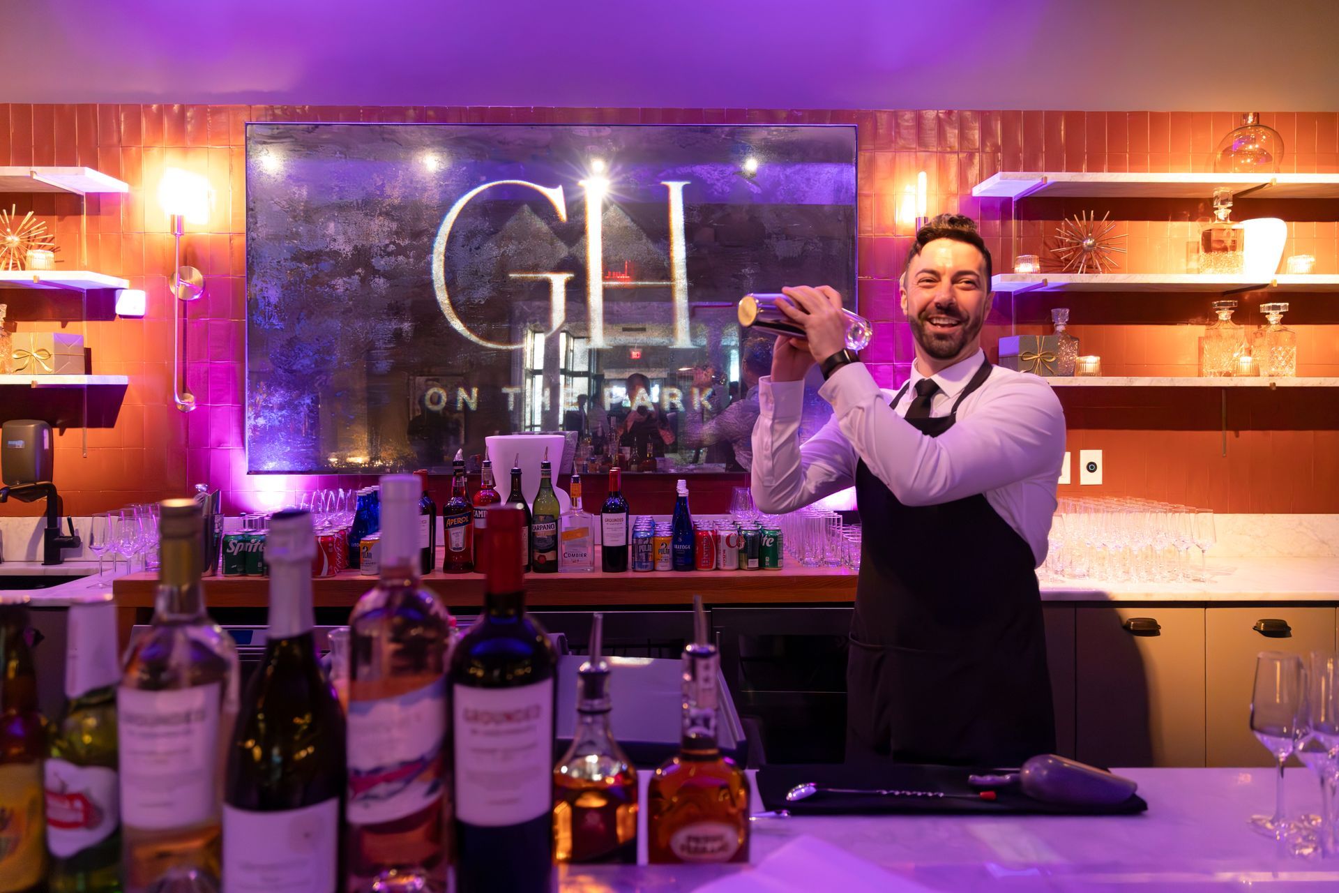Bartender shaking a cocktail in a purple-lit bar with bottles displayed on the counter.