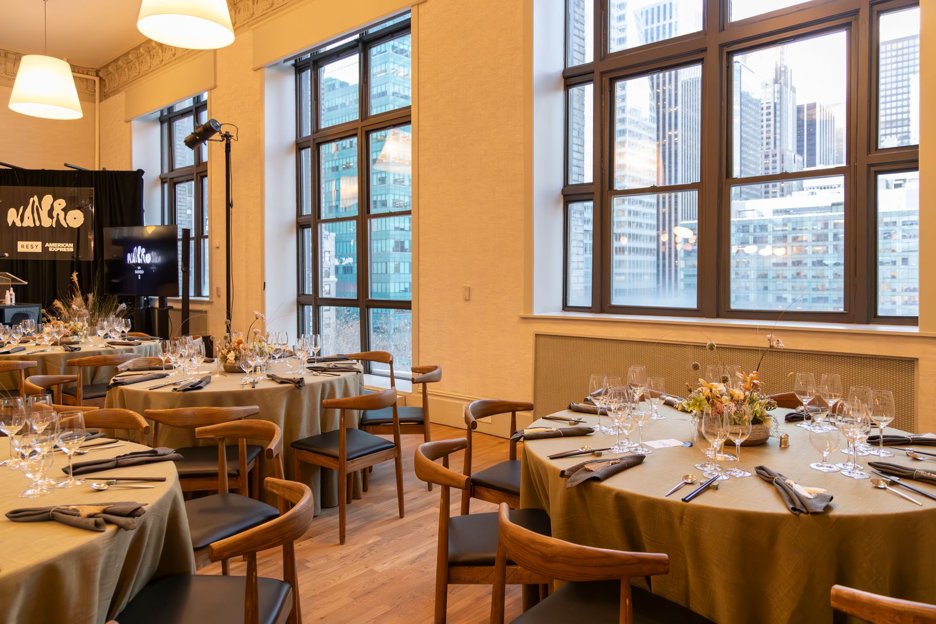 Elegant restaurant dining room with set tables, warm lighting, and large windows overlooking city buildings