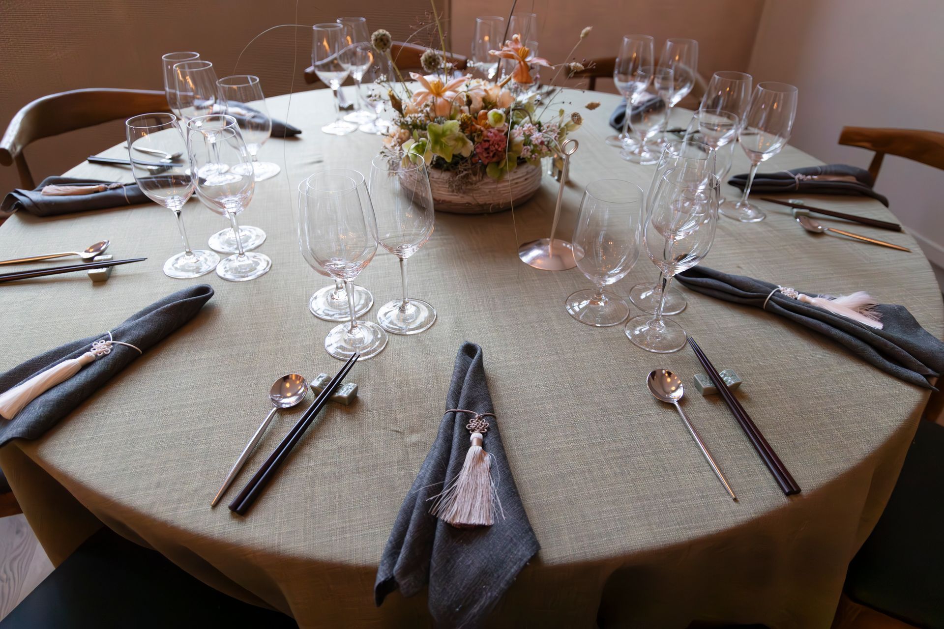 Round dining table set for a formal meal, with wine glasses, folded napkins, silverware, and a floral centerpiece.