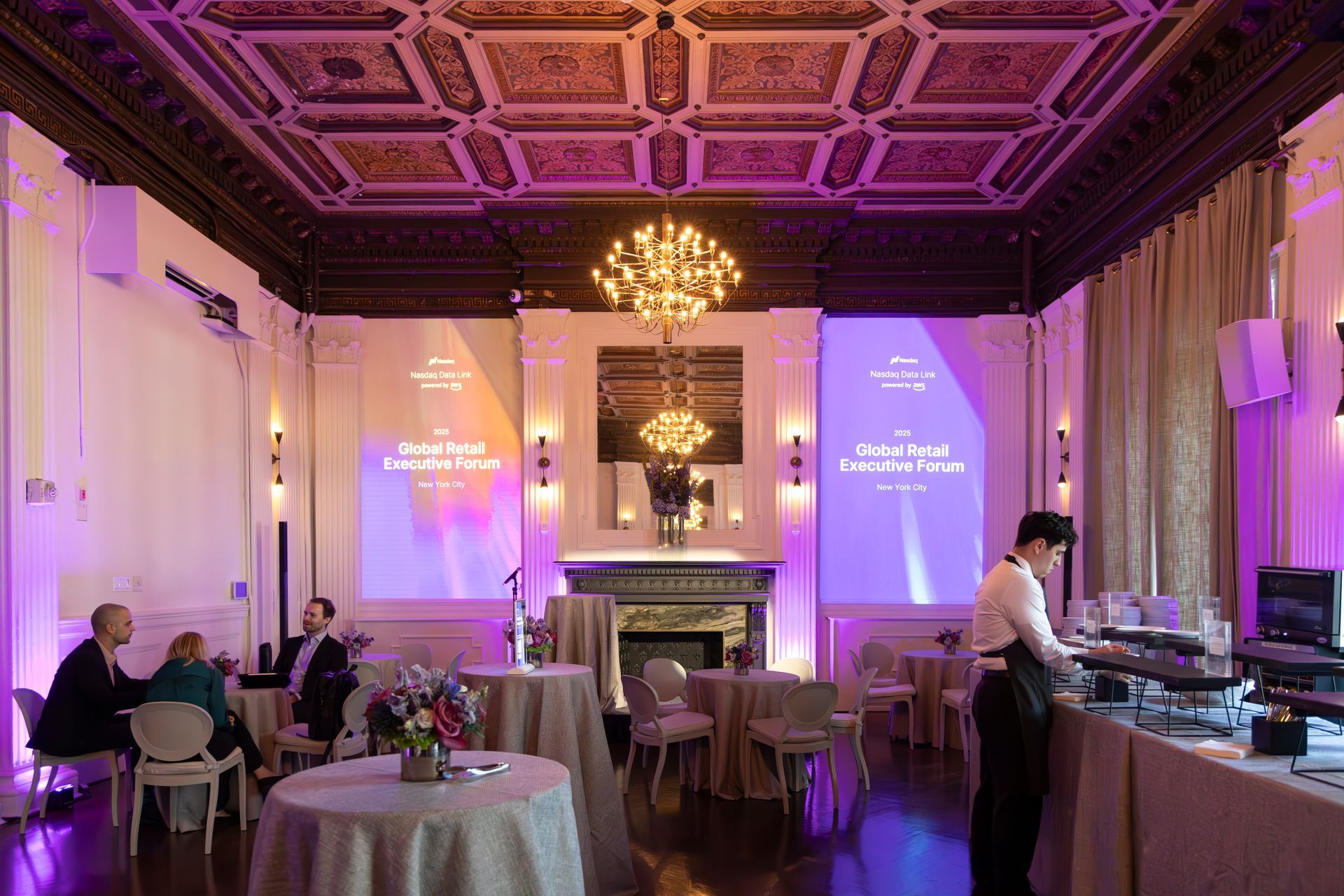 Elegant banquet hall with purple lighting, chandeliers, round tables, and a server at the buffet counter