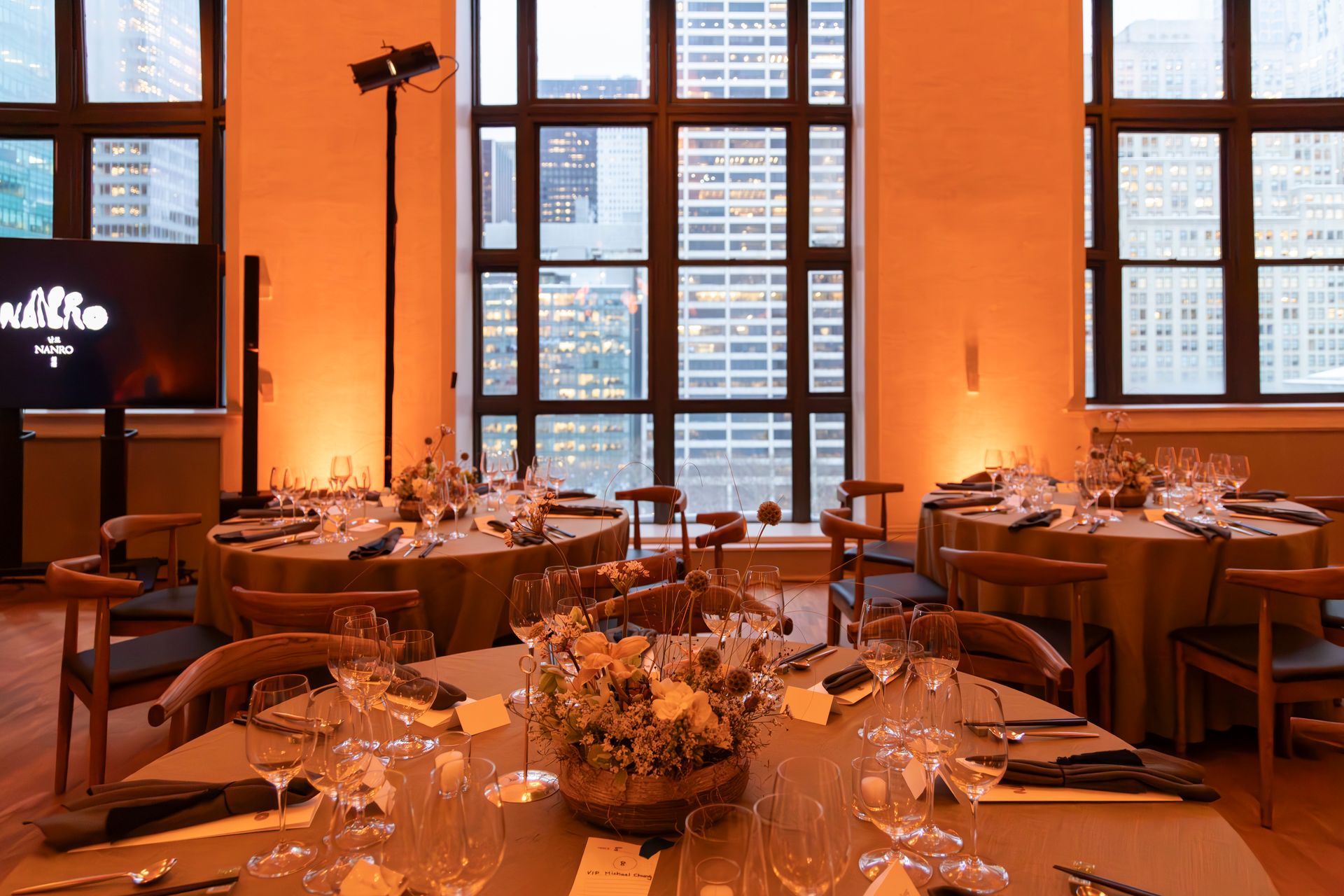 Elegant banquet hall with round tables, candles, and tall windows glowing with warm orange light.