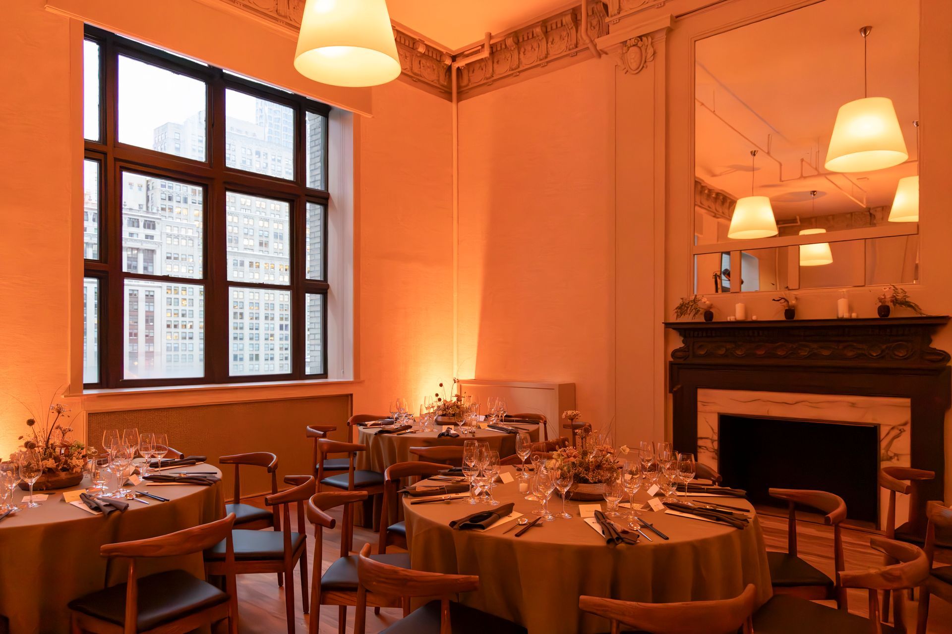 Warmly lit restaurant dining room with set tables, chairs, fireplace, and large windows at dusk