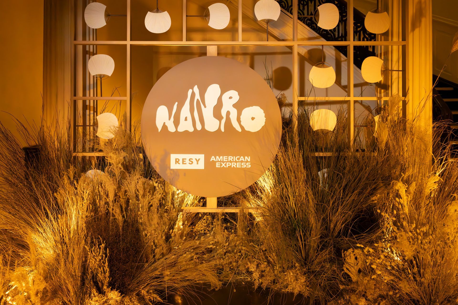 Orange-lit event display with a round NAIRO sign, glass backdrop, and tall decorative grasses outdoors