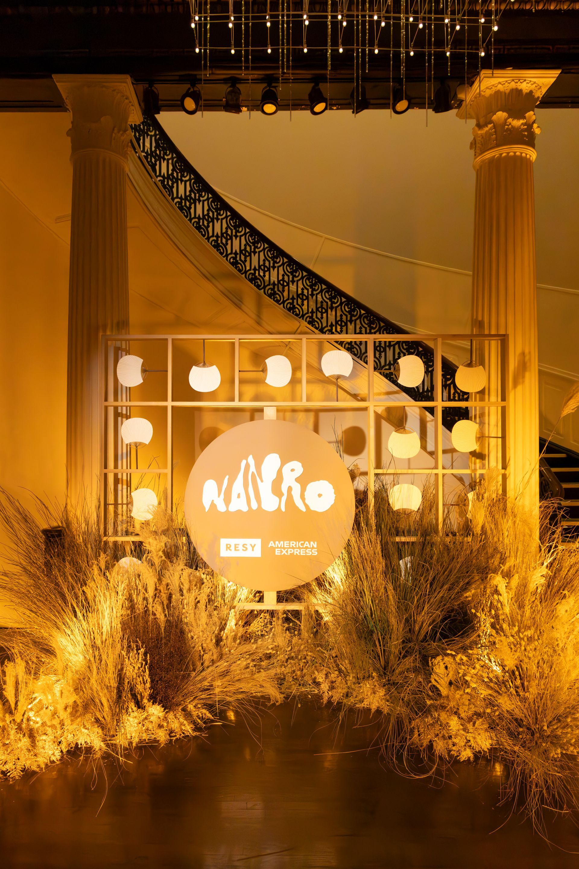 Nighttime exterior of a warmly lit building with columns and a sweeping staircase behind glowing bushes.