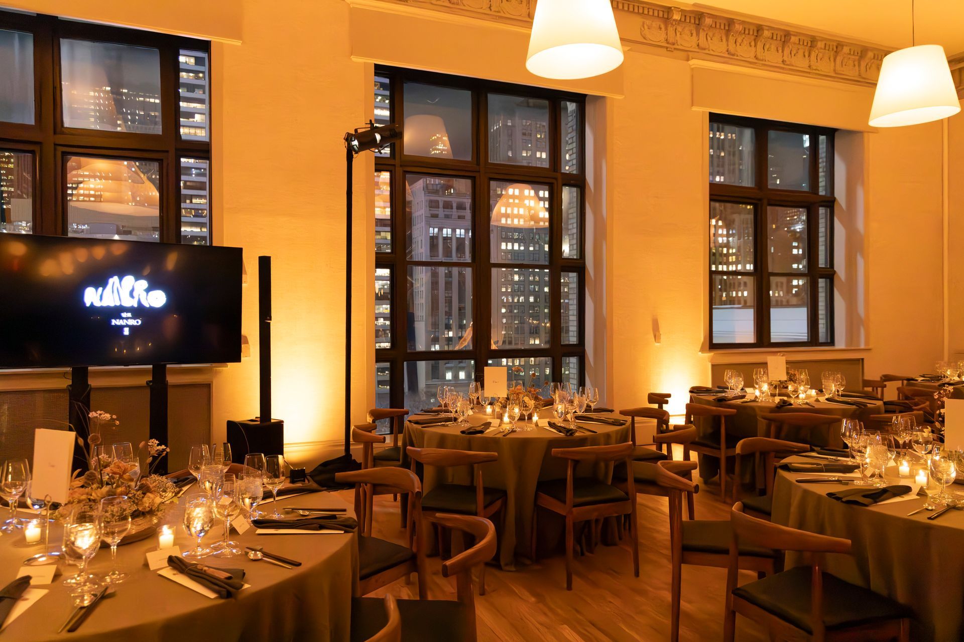 Warmly lit restaurant dining room with set tables, wooden chairs, large windows, and hanging pendant lights