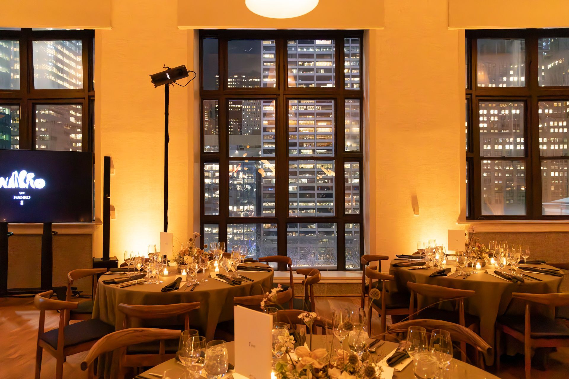 Warmly lit restaurant dining room with set tables, candles, and tall windows at dusk