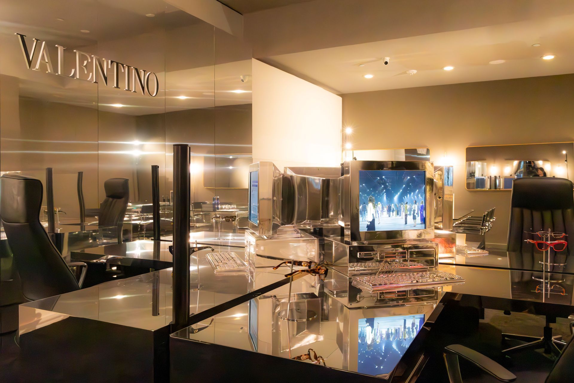 Modern showroom with glossy black tables, chrome chairs, and a “VALENTINO” sign reflected in mirrors.