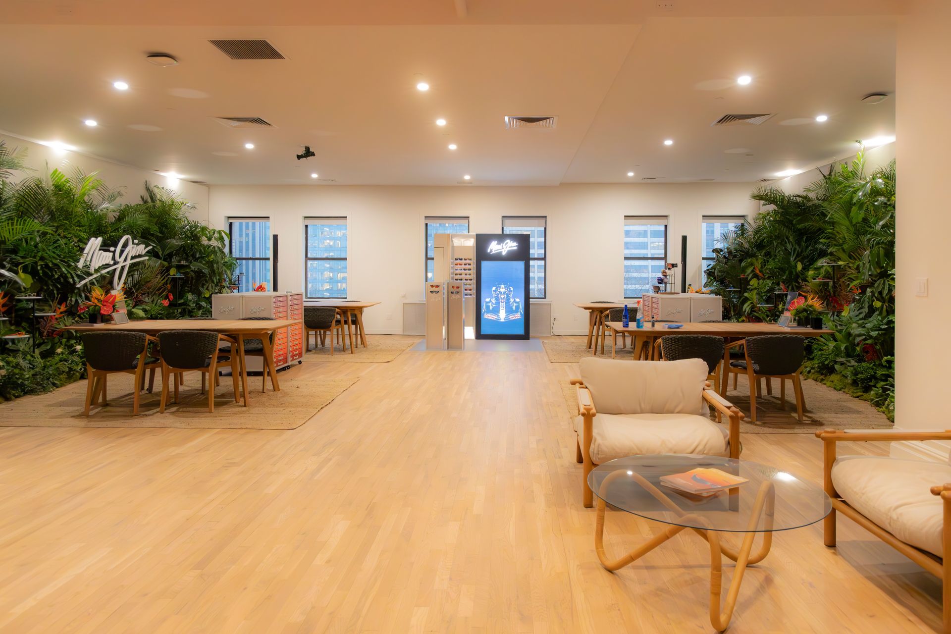 Bright, modern lounge with wood floors, indoor plants, tables, and chairs under warm ceiling lights.