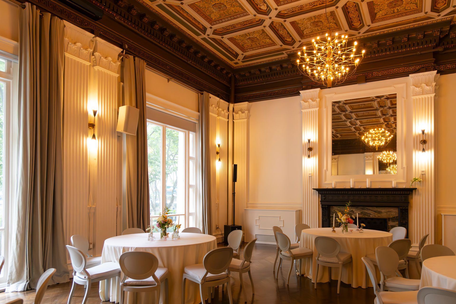 Elegant dining room with chandeliers, ornate ceiling, tall windows, and set tables with white tablecloths.