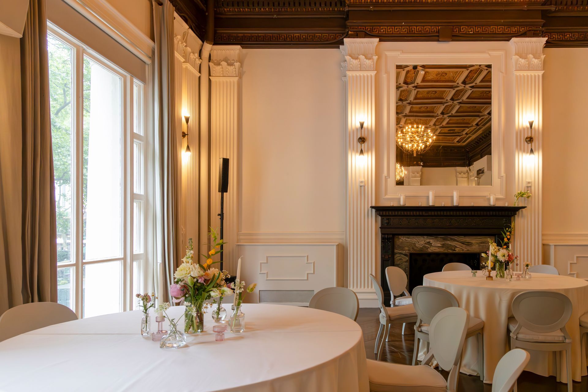Elegant dining room with round tables, white chairs, flowers, and a fireplace beneath a gold-framed mirror
