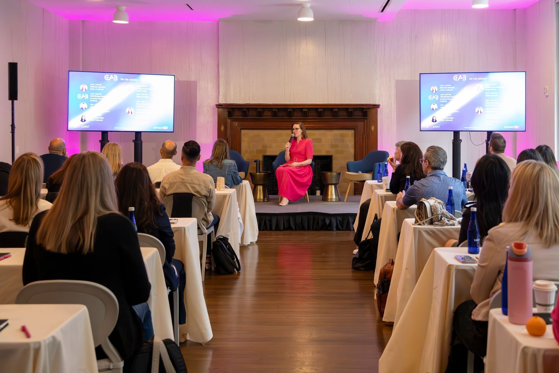 Conference audience facing a speaker on a small stage with two presentation screens and pink lighting