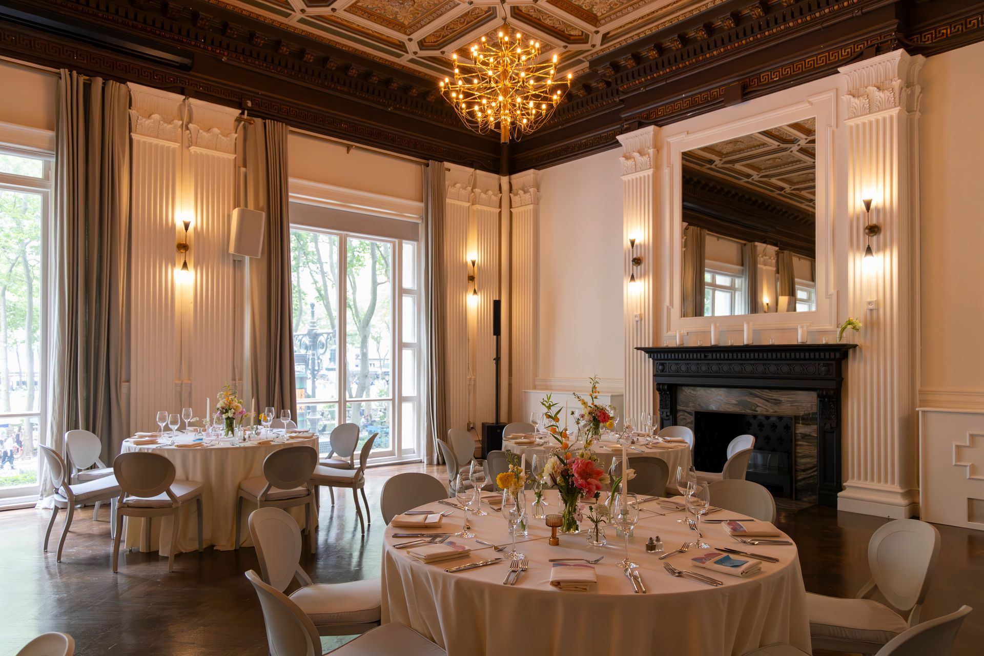 Elegant dining room with round tables set for a formal event, tall windows, chandeliers, and a marble fireplace