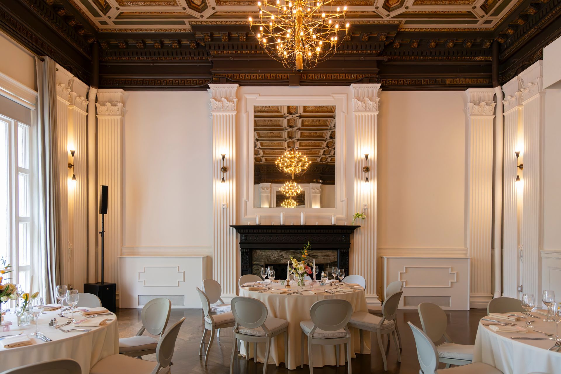 Elegant dining room with chandeliers, ornate fireplace, and set tables in a bright formal hall