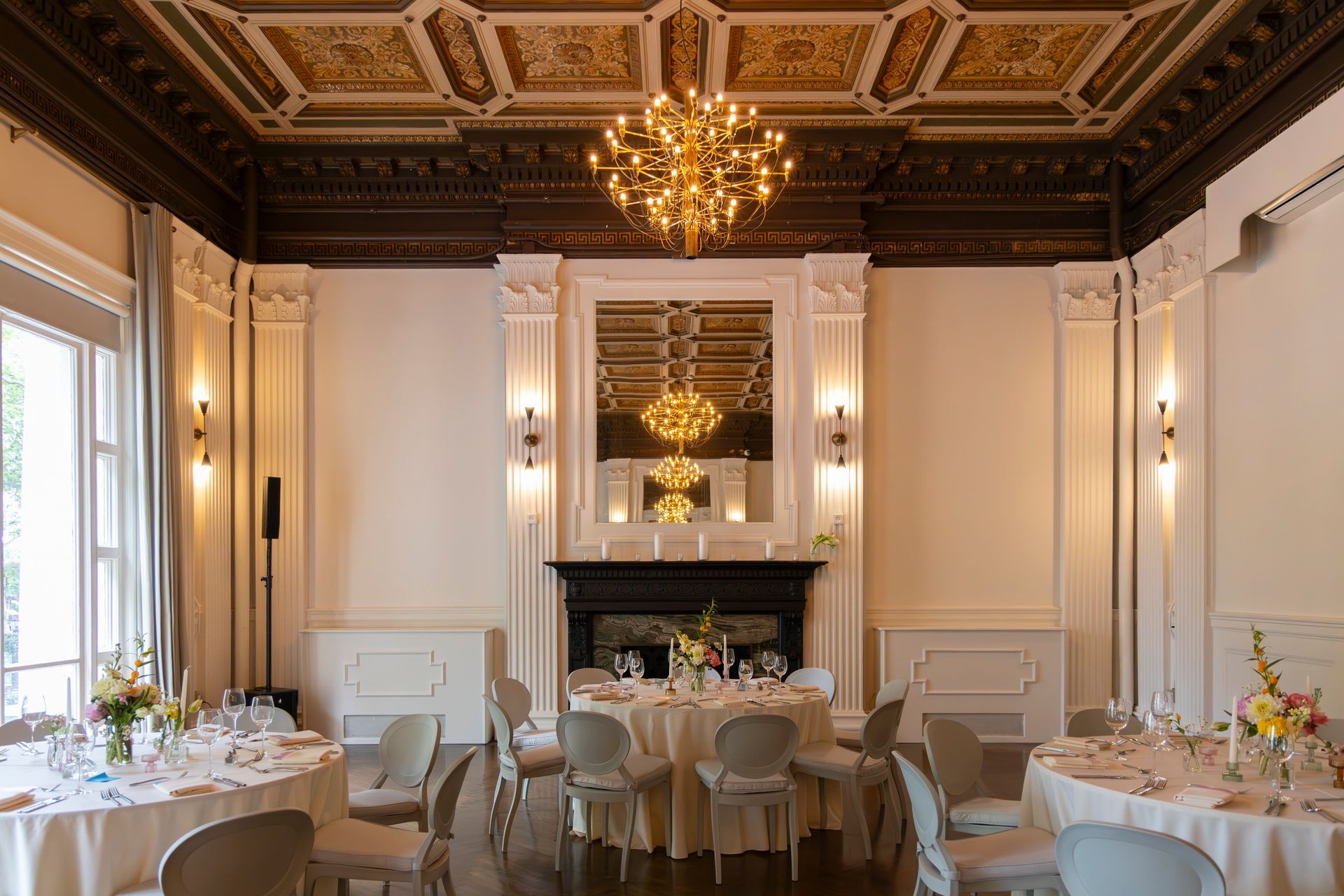 Elegant formal dining room with chandelier, ornate ceiling, white chairs, and set tables by tall windows