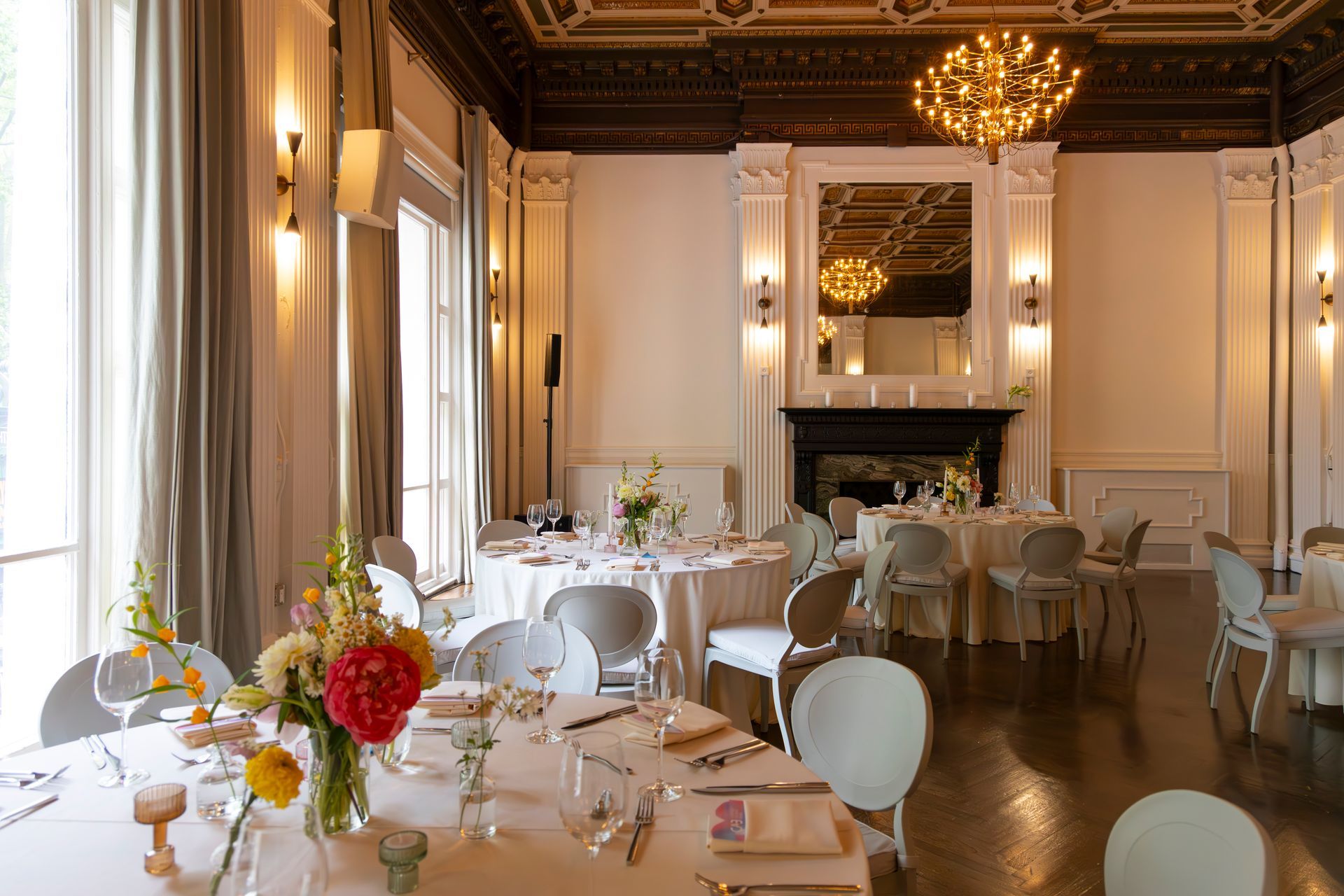 Elegant dining room with set tables, white chairs, chandeliers, and a large fireplace.