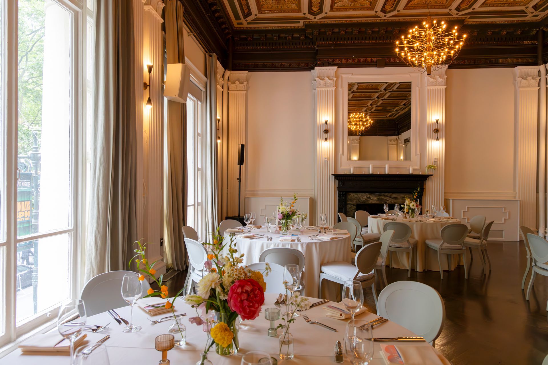 Elegant dining room with set tables, white chairs, chandeliers, and a fireplace