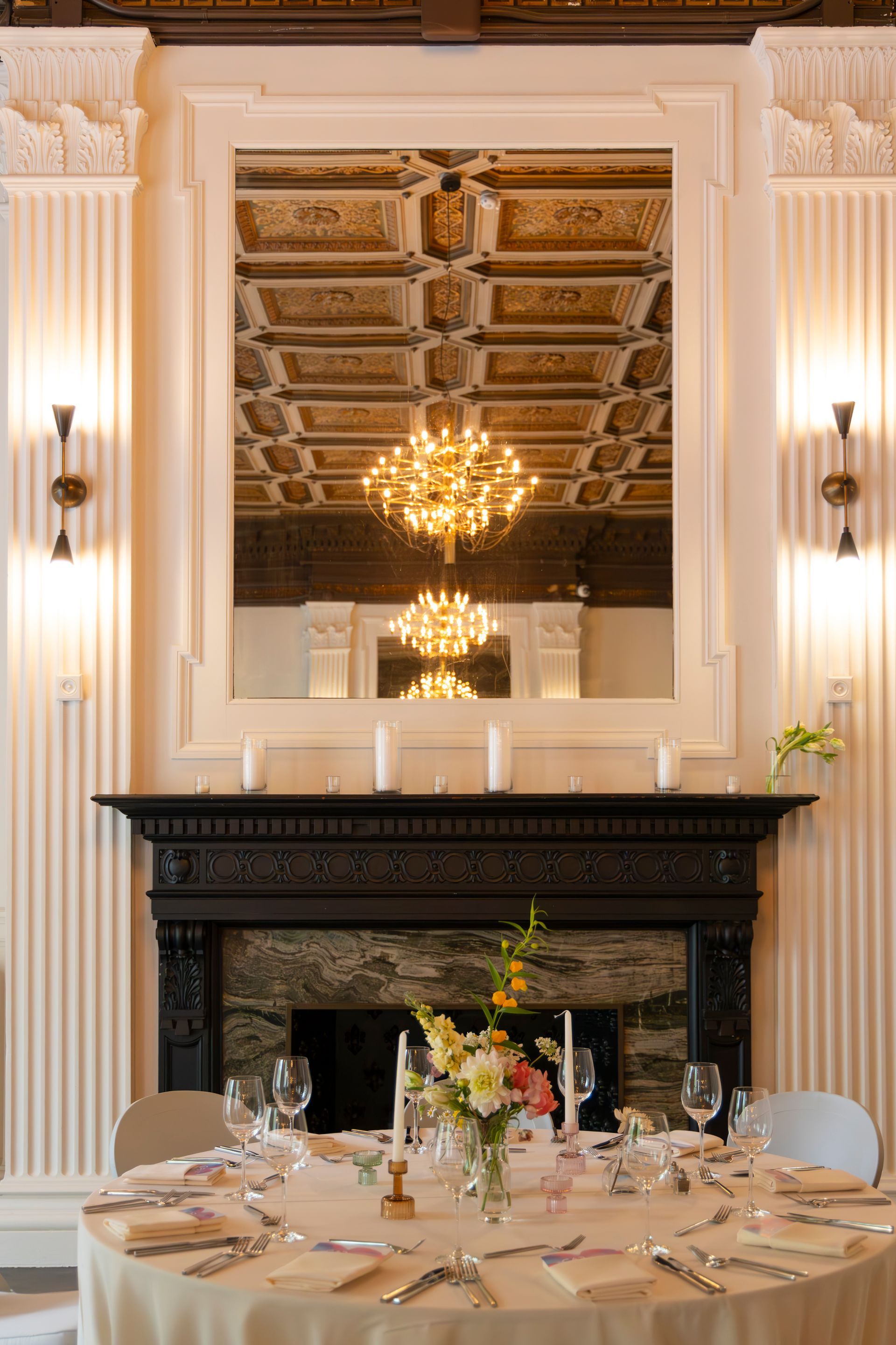Elegant dining room with set round table, black fireplace, chandeliers, and ornate ceiling.