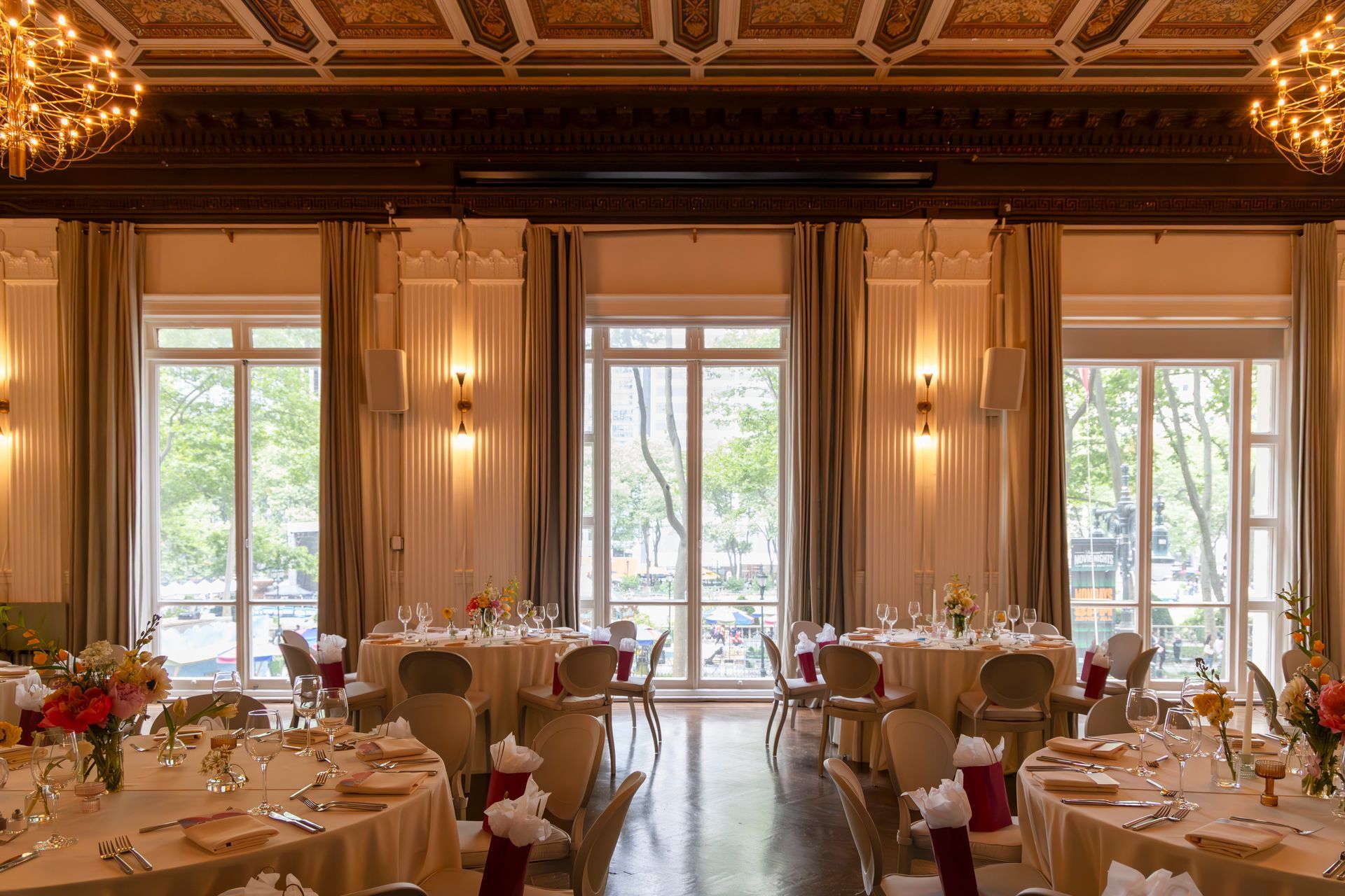 Elegant banquet hall with round tables, chandeliers, and tall windows overlooking greenery