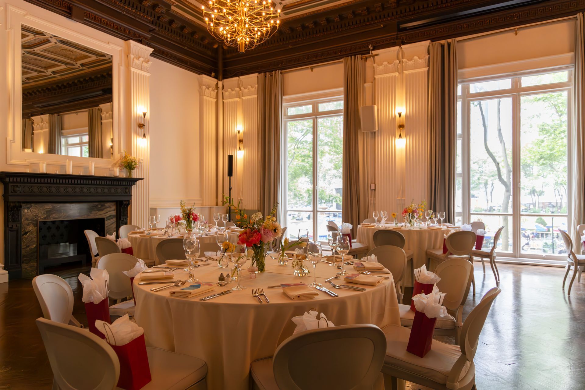 Elegant dining room with round tables, white linens, chandeliers, and large windows overlooking a garden.
