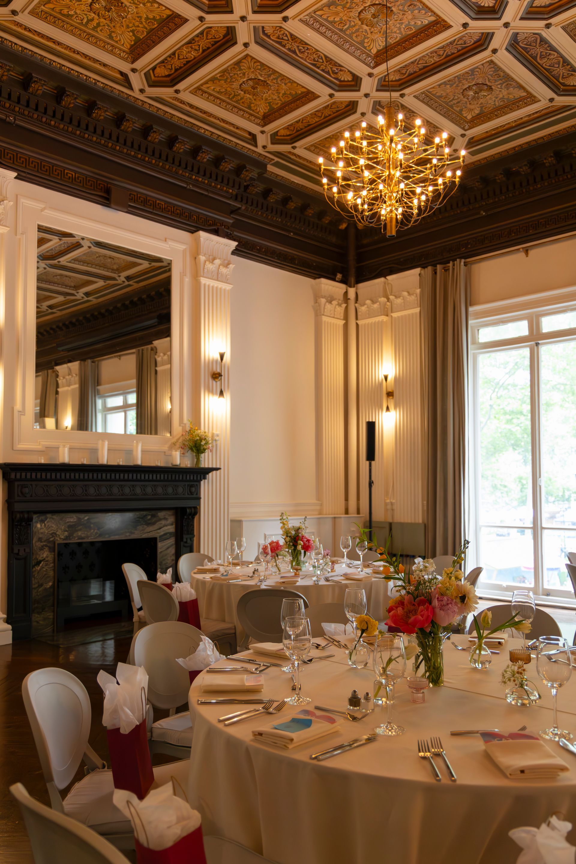 Elegant dining room with ornate ceiling, chandelier, fireplace, and set round tables by tall windows