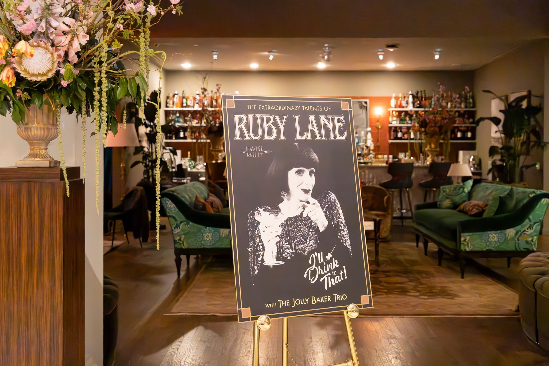 Ruby Lane sign on an easel in a warmly lit lounge with green velvet sofas and floral decor