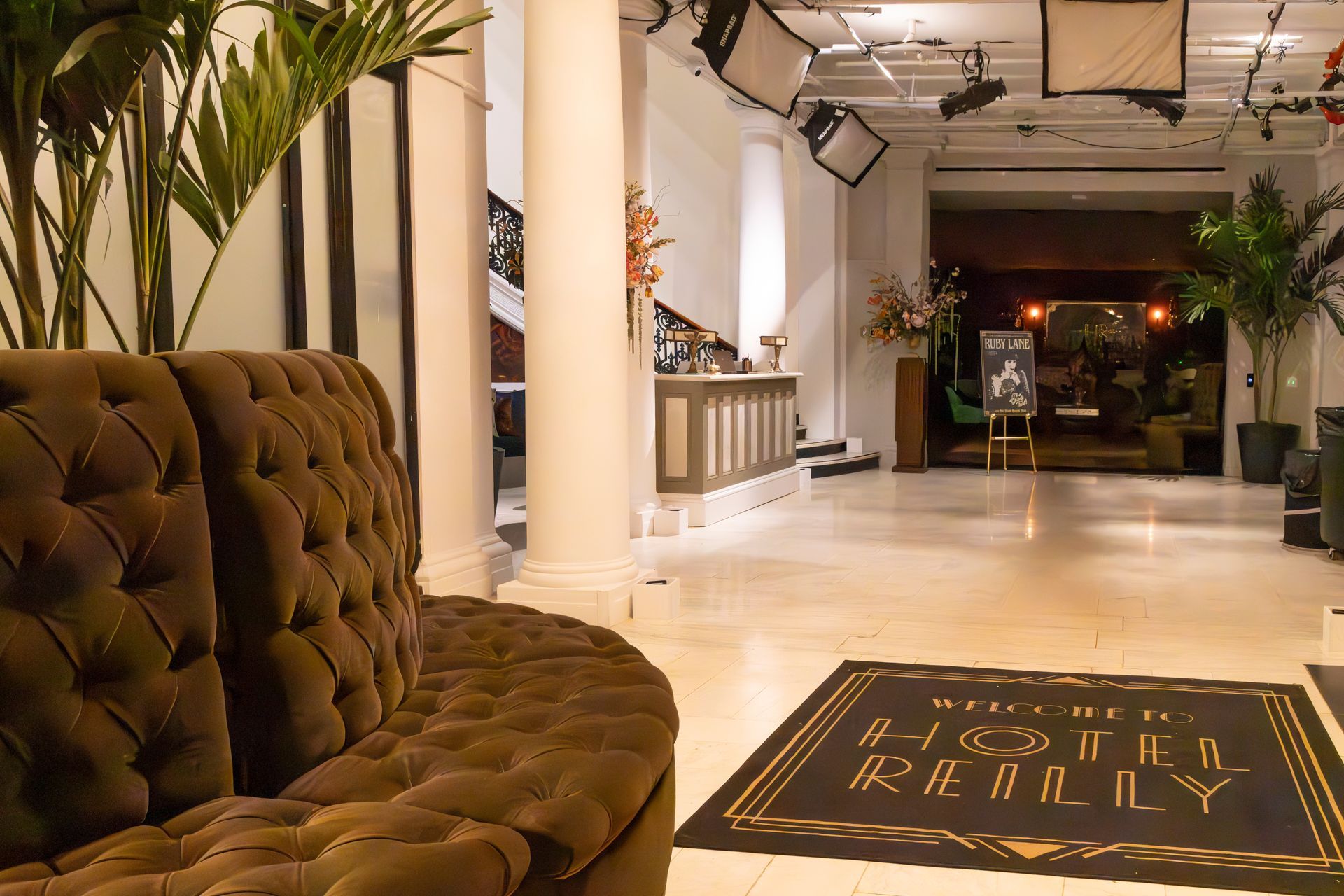Elegant hotel lobby with tufted brown sofa, white columns, and a black floor mat reading “Hotel Beverly”