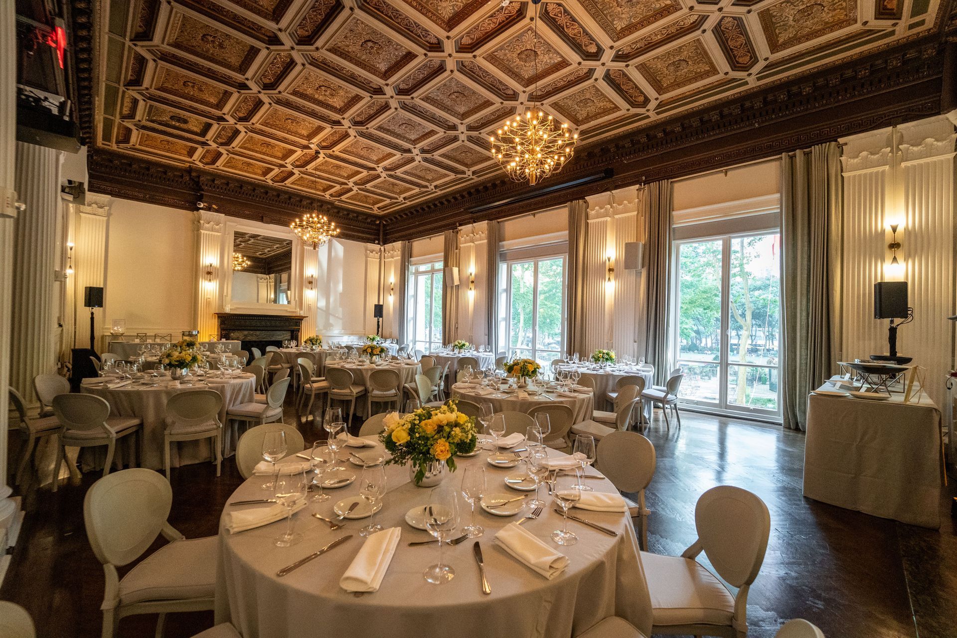 Elegant banquet room with round, set dining tables, chandeliers, and large windows with sheer curtains