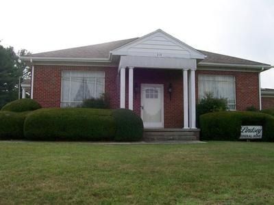 A one-story brick house with a front porch, white pillars, a central white door, and large, well-trimmed bushes out front.