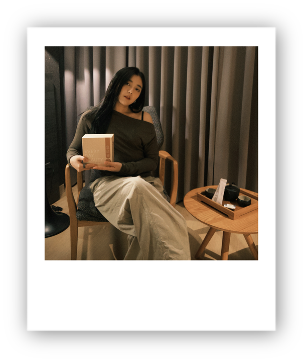 Woman sitting in a chair, holding a box, with a table and curtains in the background.