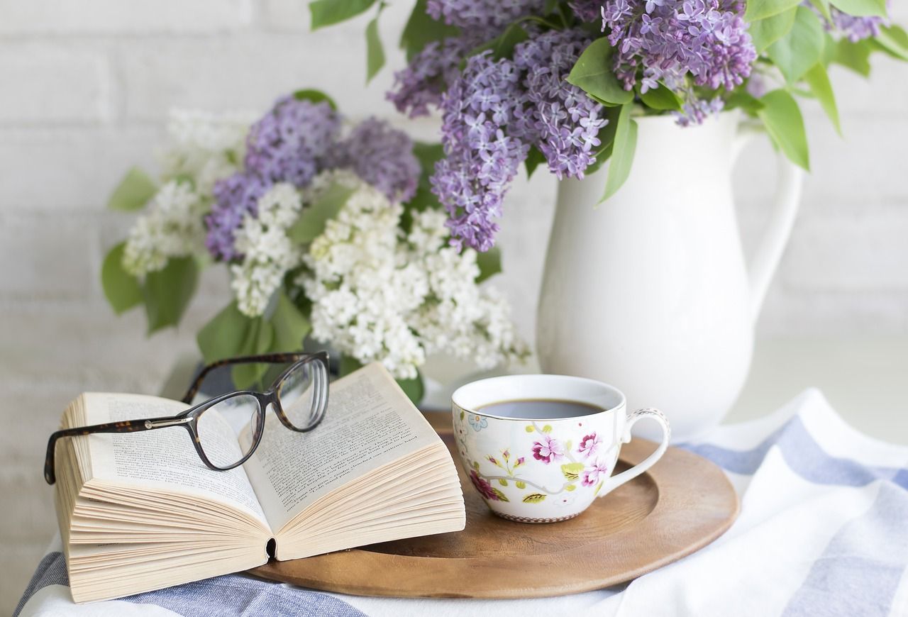 Open book with glasses, floral teacup, wooden tray, and lilac bouquet on a table.