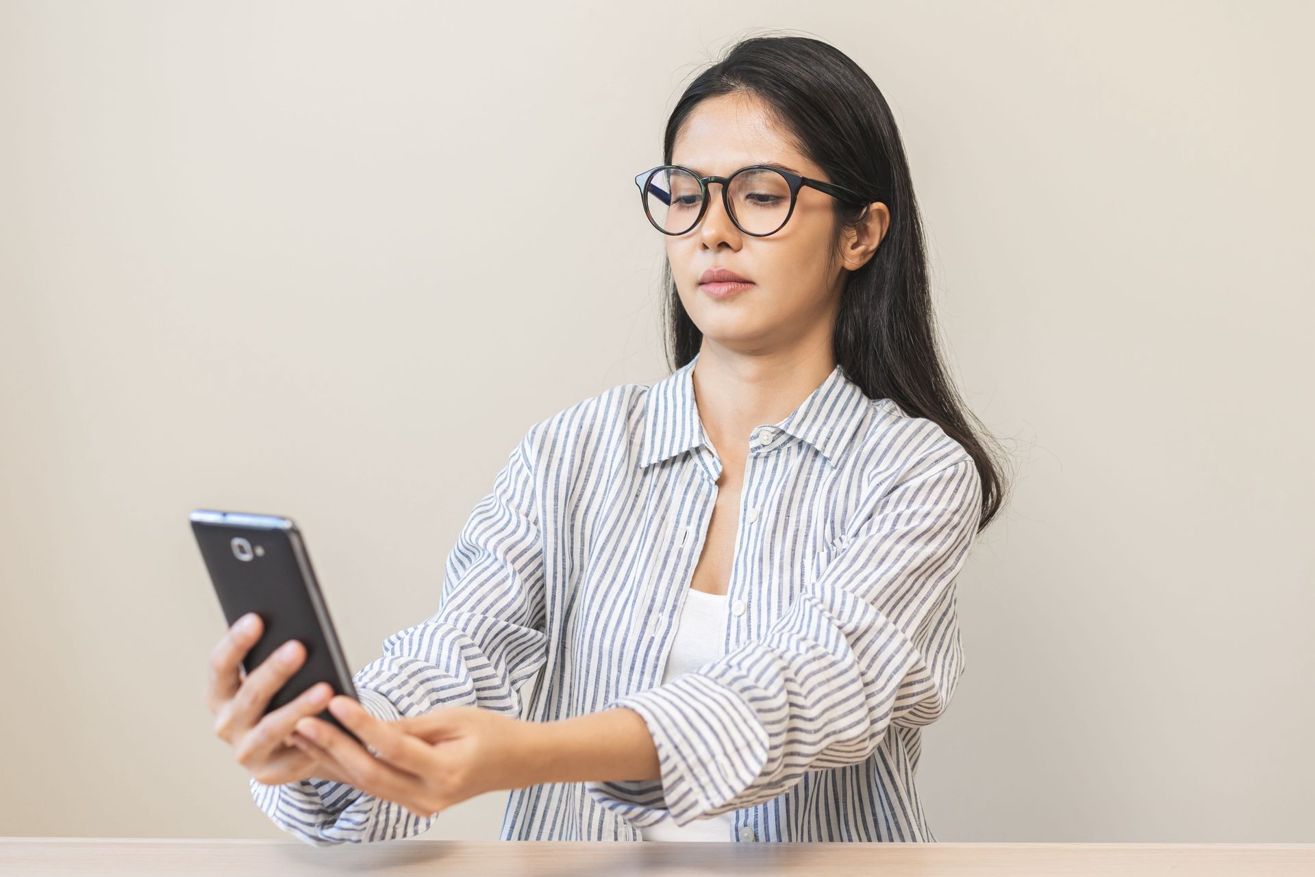 Woman wearing glasses, looking at a mobile phone while seated at a table. She wears a striped shirt.