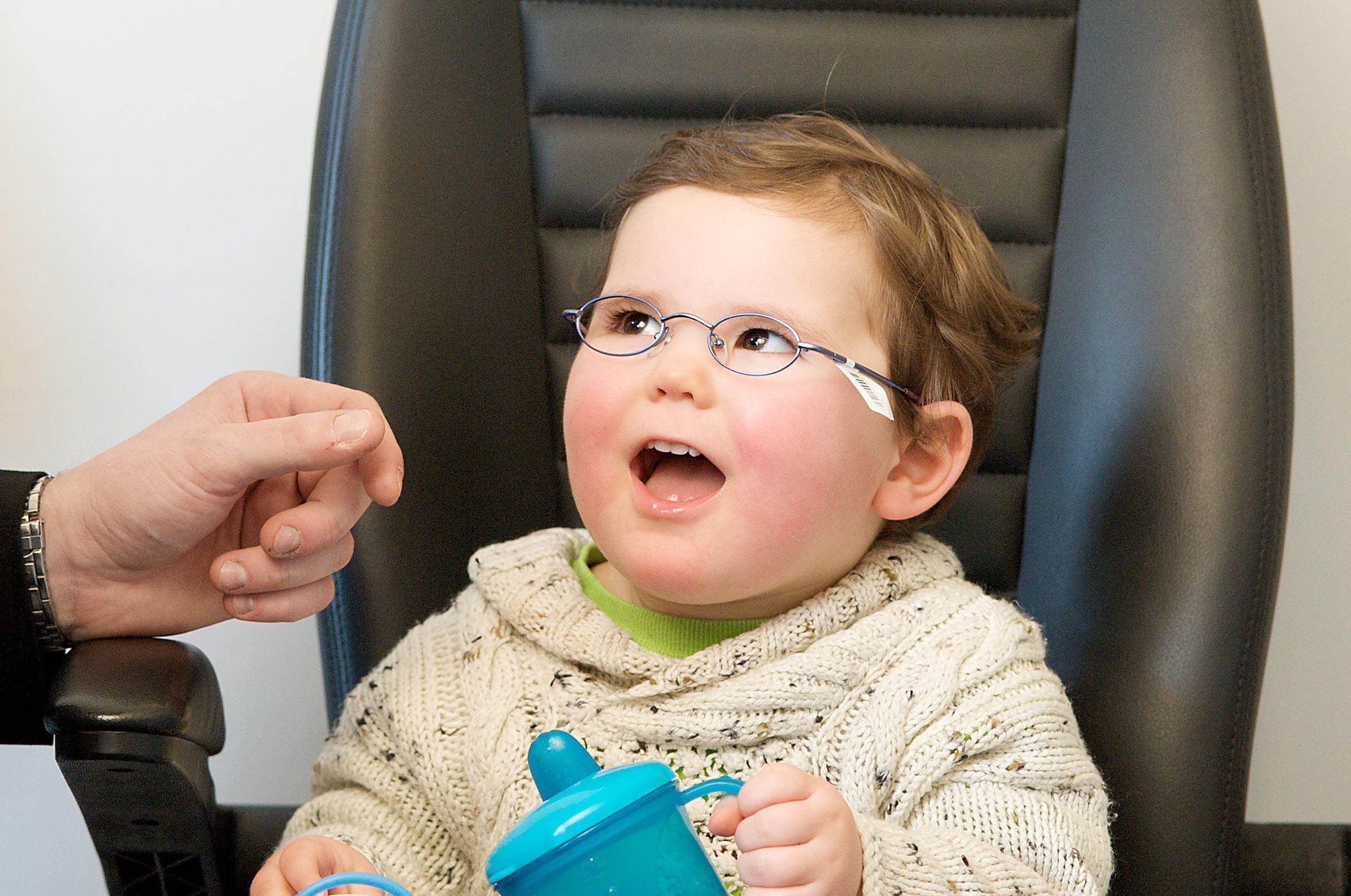 Child wearing glasses, seated in a chair, with mouth open and holding a sippy cup. Someone's hand is near them.