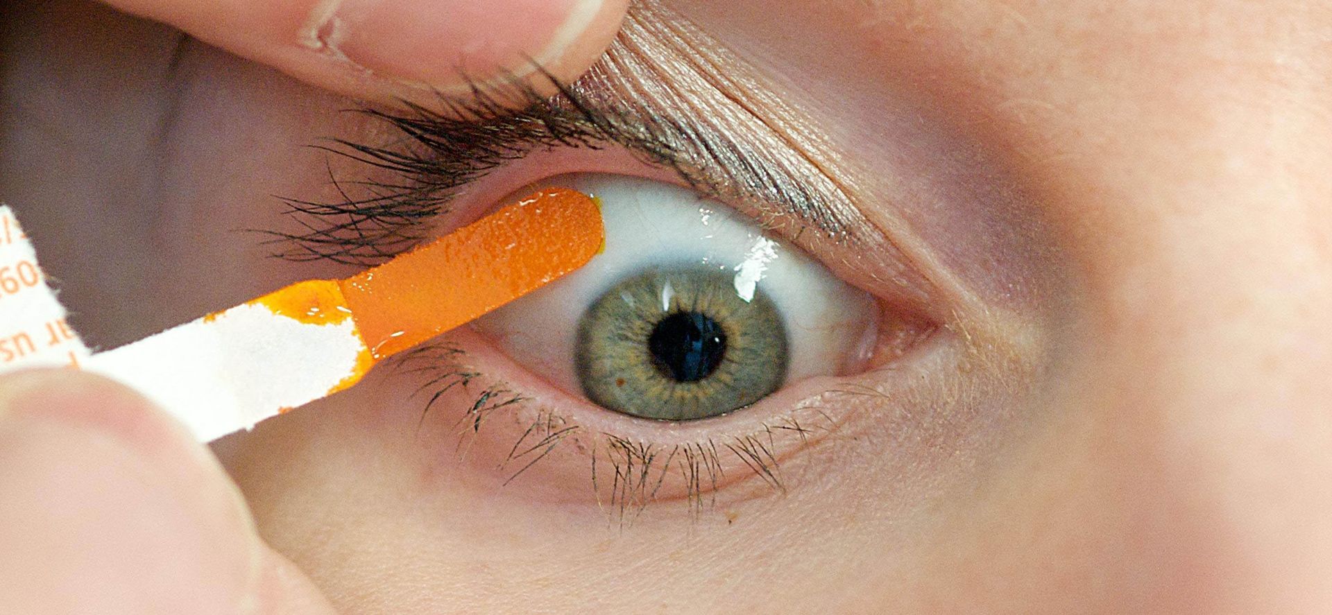 An eye being tested with an orange strip. The eye is open, with a green iris. A hand is holding the strip.