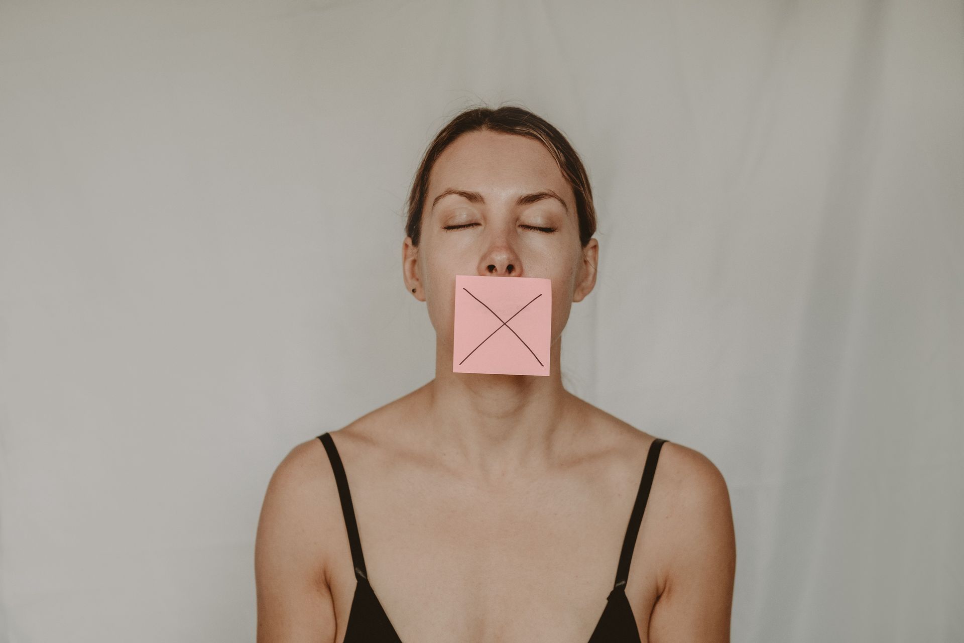 A woman in a black top closes her eyes and has her mouth covered with a Post-It displaying a black X to hide her mouth.