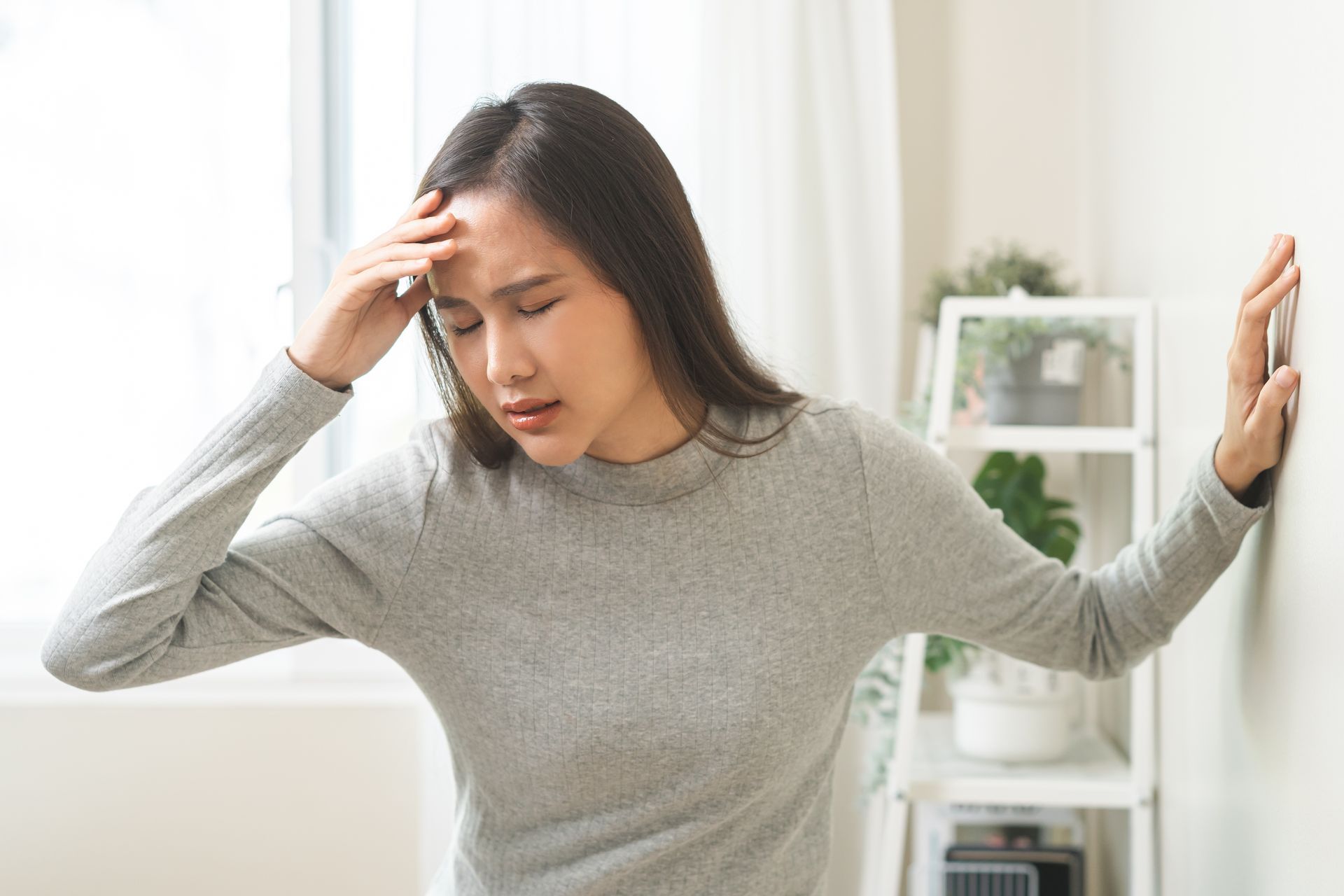 Young Asian woman in a grey sweatshirt holds the wall while suffering from dizziness due to TMD symptoms.