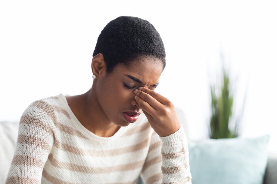 African American woman in a striped cardigan holds the top of her nose from sinus pain.