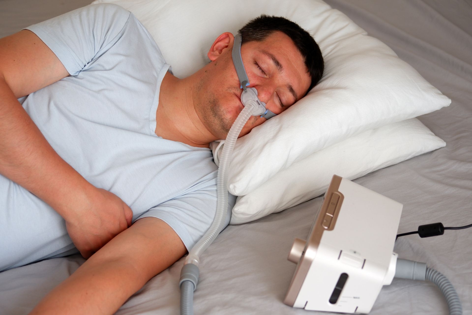 Middle aged man in white t-shirt laying in bed with a CPAP mask on while sleeping.