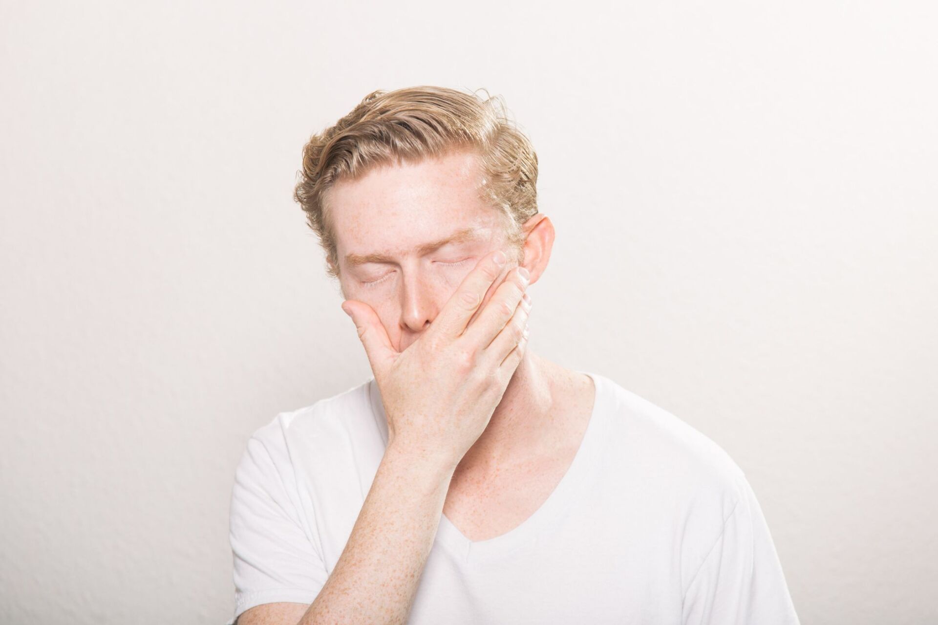 Middle aged man in white t-shirt holds hand over mouth while feeling pain from grinding his teeth.
