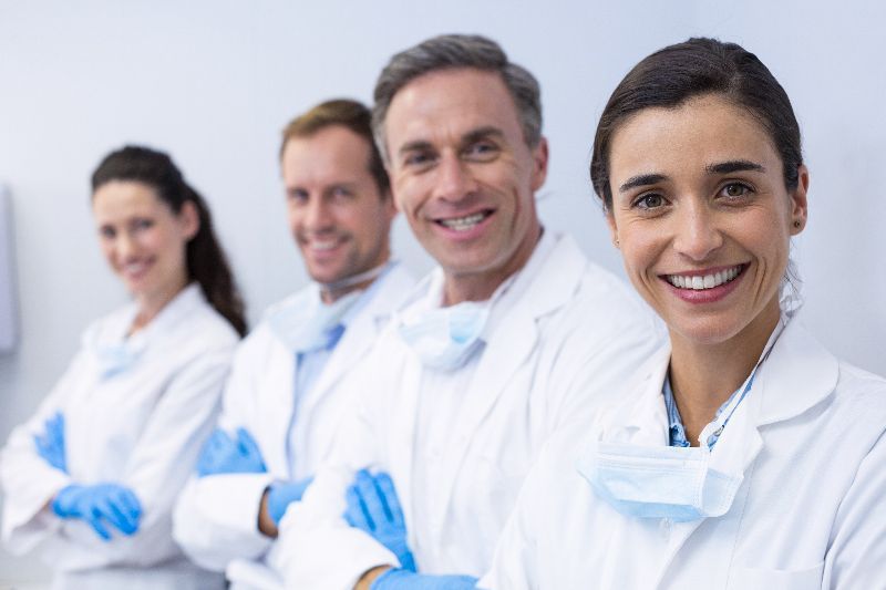 Four doctors in white scrubs with blue gloves smile for the camera against a white background.