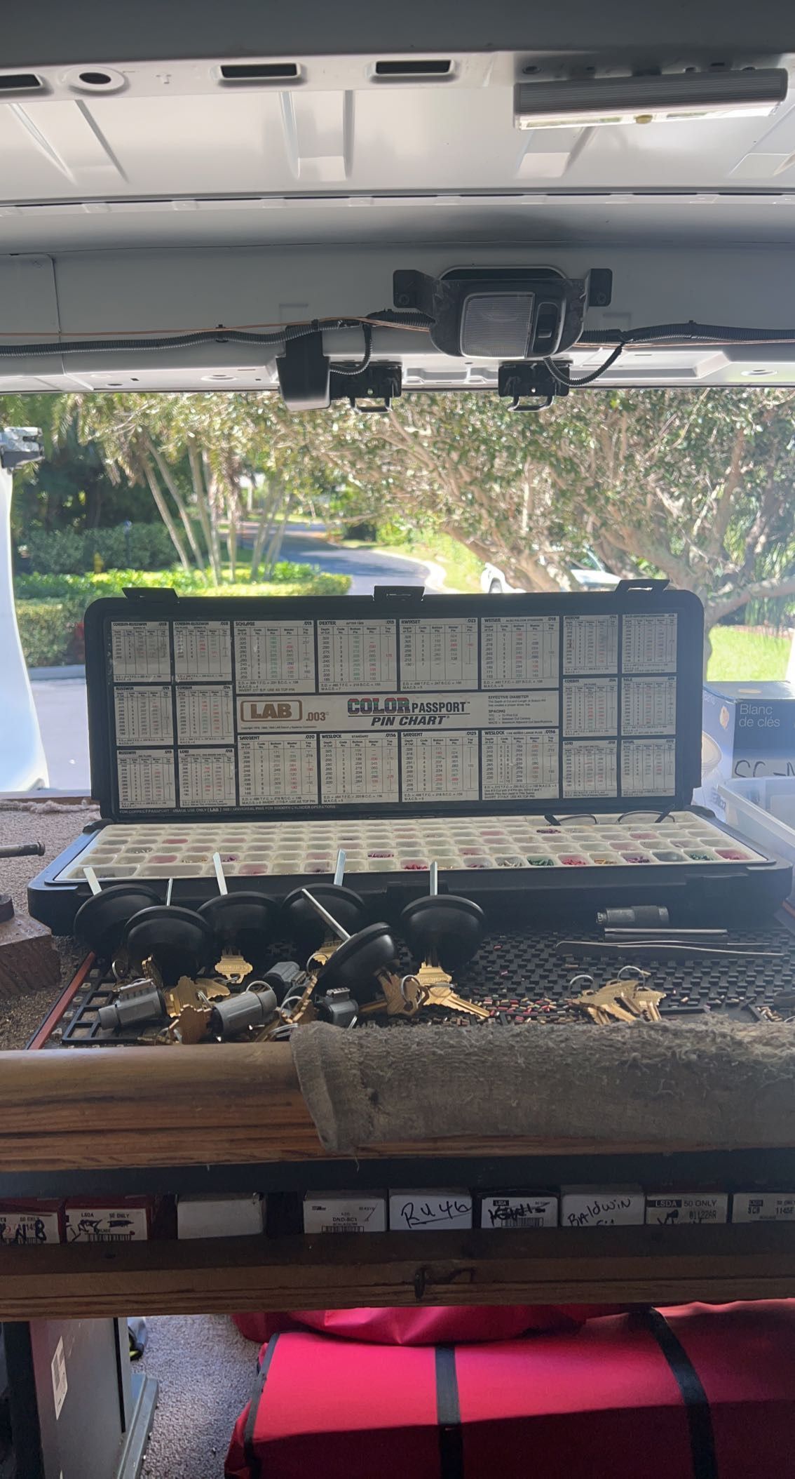 Open tackle box with assorted fishing gear on a vehicle dashboard, parked near a green wooded area