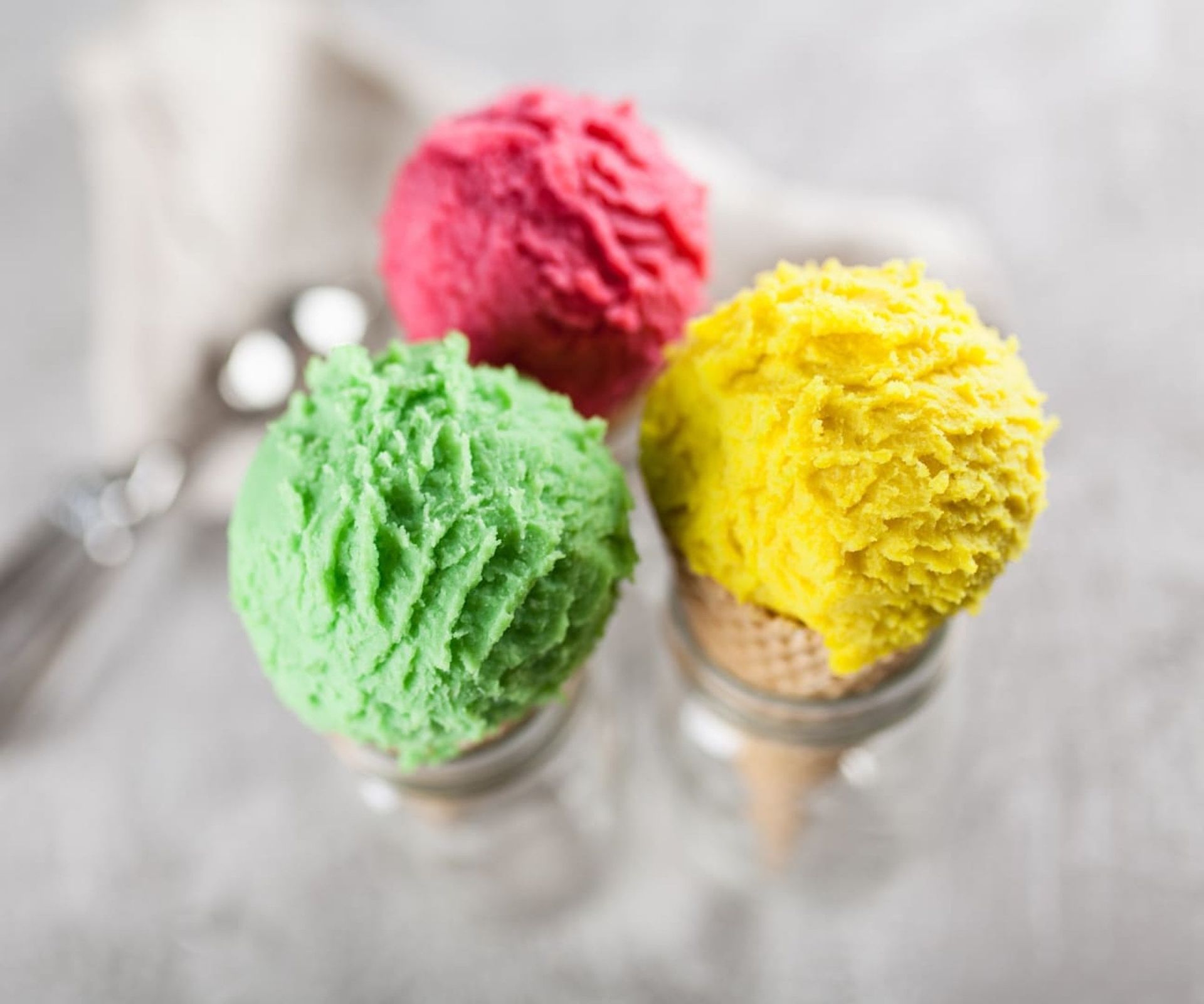 Three scoops of colorful ice cream: green, pink, and yellow, in glass jars with a silver scoop in the background.
