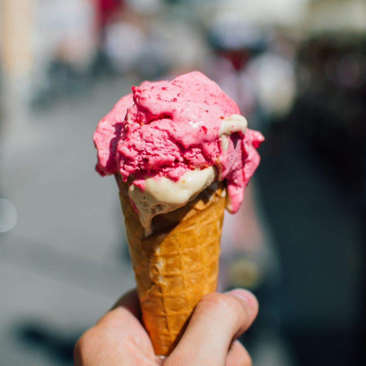 Hand holding a waffle cone with two scoops of melting pink ice cream. Blurry outdoor background.