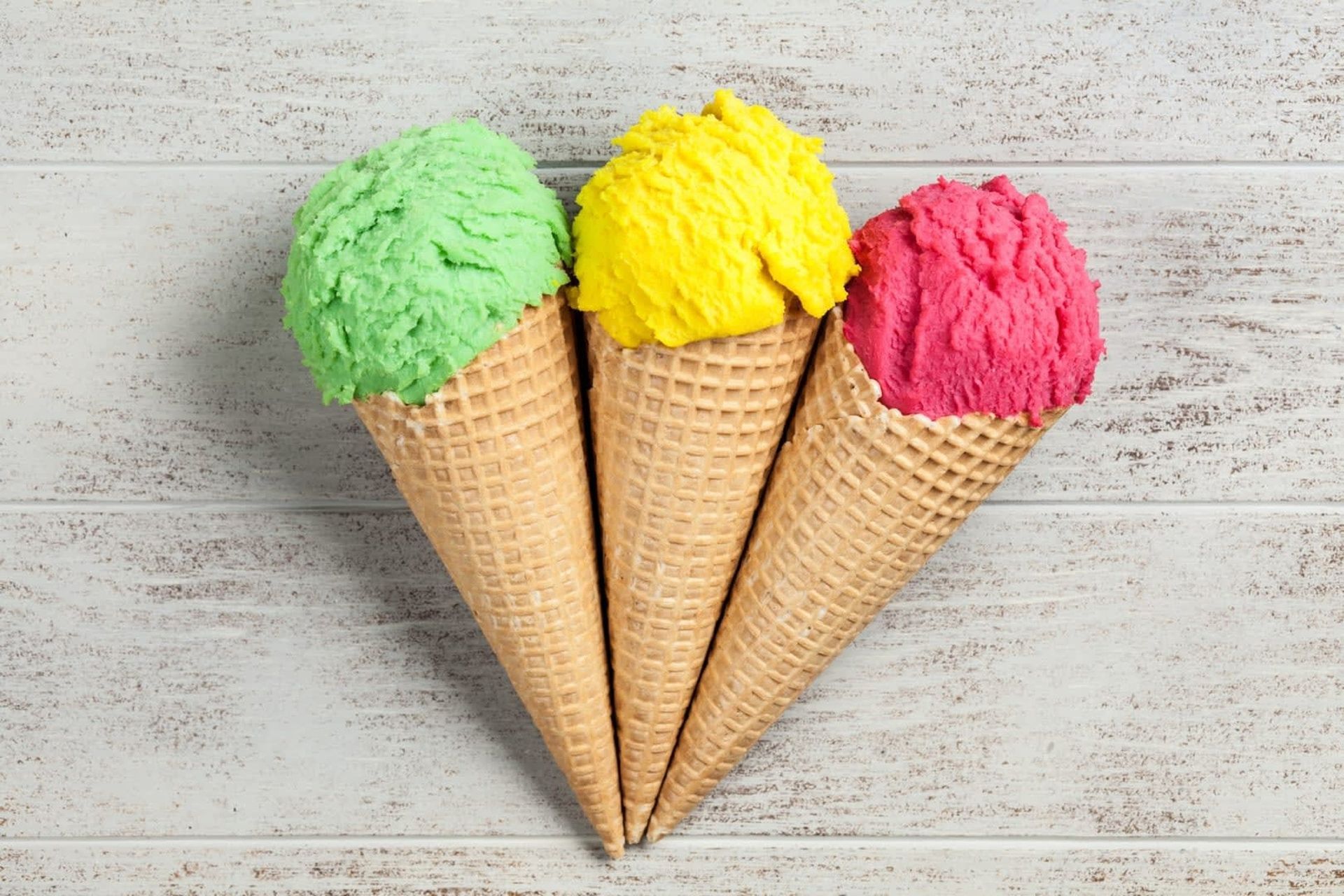 Three ice cream cones: green, yellow, and pink scoops in waffle cones, set against a white wood-grain backdrop.