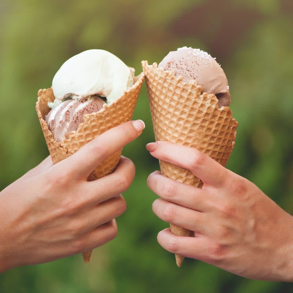 Two hands holding ice cream cones, toasting. Vanilla and chocolate ice cream against a green background.