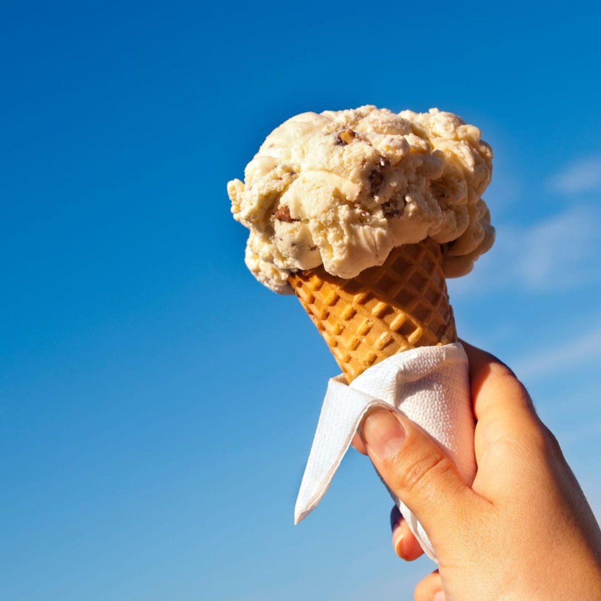 Hand holding a waffle cone with two scoops of ice cream against a blue sky.