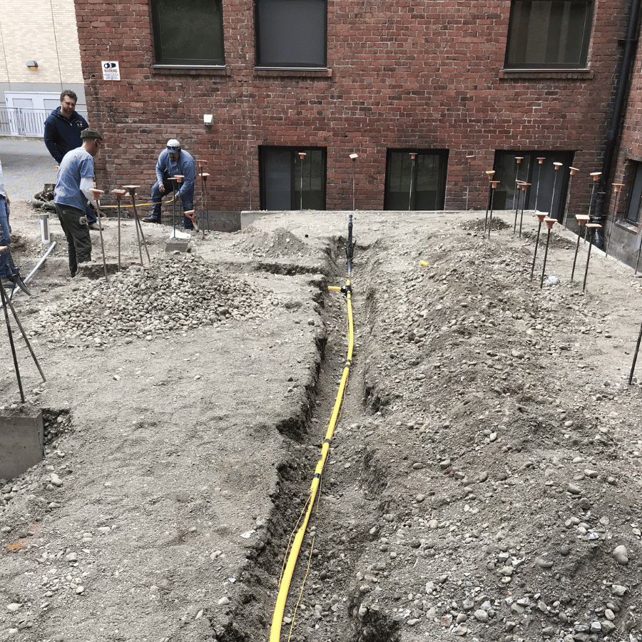Construction workers on a gravel site with a yellow hose trench beside a brick building.