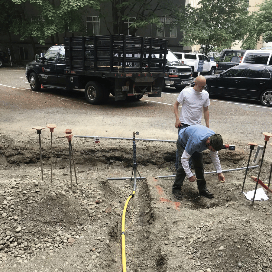 Two workers digging a trench beside parked trucks in a gravel construction area