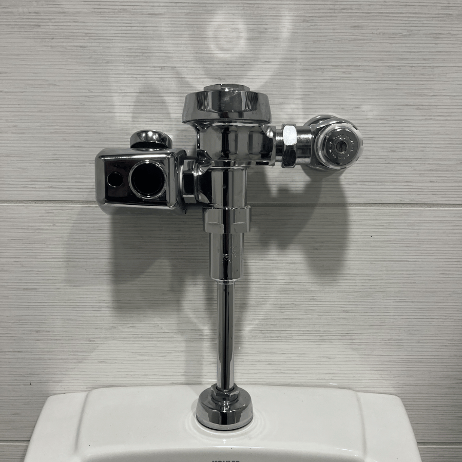A chrome urinal flush valve mounted on a tiled wall, casting a shadow.