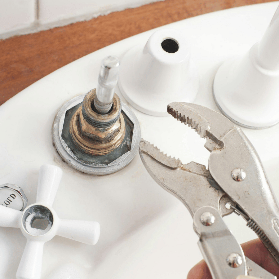 Pliers grip a faucet stem on a white sink, with a detached handle nearby, indicating plumbing repair in progress.