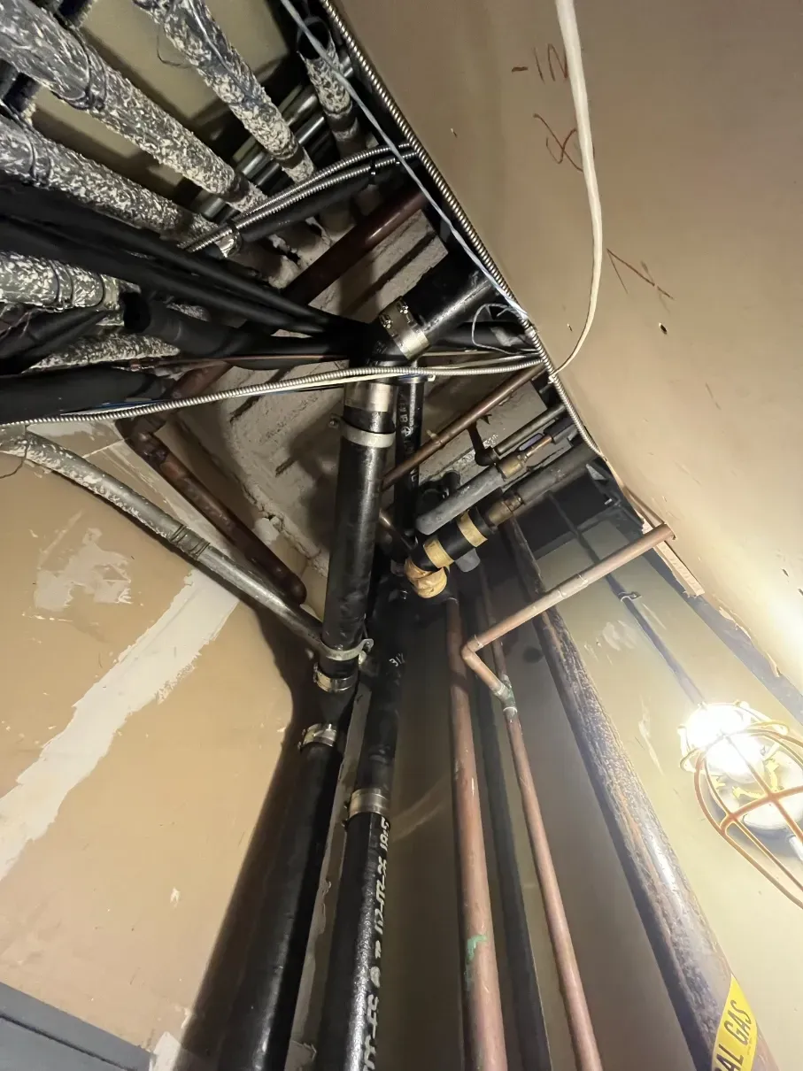 Industrial ceiling with exposed black pipes and wiring in a beige utility corridor